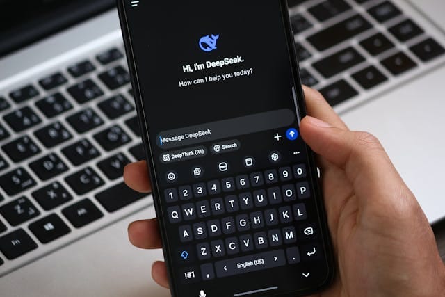 Zbliżenie na dłoń trzymającą smartfon z włączoną aplikacją czatu o nazwie "DeepSeek", gotową do wpisania wiadomości, na tle klawiatury laptopa. Aplikacja wykorzystywana jest do głębokiego przeszukiwania informacji, co rodzi pytania, czy klienci ufają AI skoro muszą sami weryfikować treści.