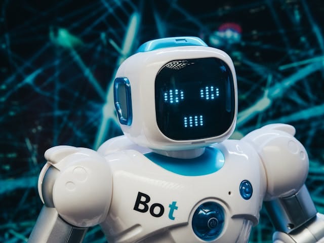 Zbliżenie na małego, białego robota o zaokrąglonych kształtach, z napisem "Bot" na klatce piersiowej. Jego cyfrowa twarz wyświetla emotikon z niebieskich kropek na tle abstrakcyjnej, technologicznej sieci. Zdjęcie odnosi się do tworzenia treści przez AI i wykorzystywaniu sztucznej inteligencji w marketingu AI w firmie Contentologia.