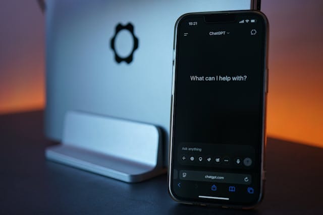 Smartfon z włączoną aplikacją do generowania treści AI ChatGPT z pytaniem "What can I help with?" na ekranie, umieszczony przed odwróconym laptopem w słabo oświetlonym, ciepłym otoczeniu. 