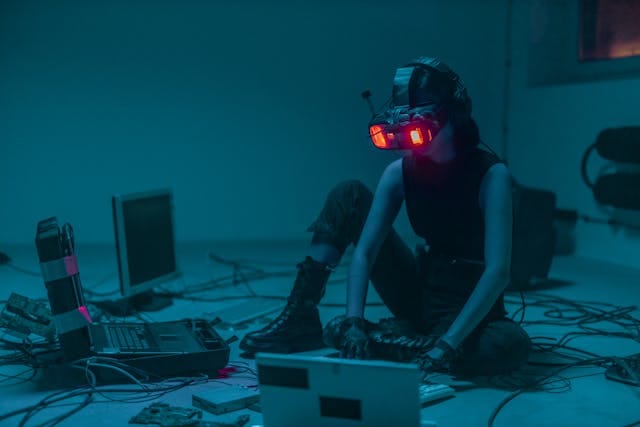Kobieta w ciemnym, futurystycznym stroju i goglach VR o czerwonych podświetlanych soczewkach siedzi na podłodze, pracując na laptopach i otoczona plątaniną kabli i sprzętu komputerowego, w klimacie cyberpunka. Zdjęcie symbolizuje obawy o bezpieczeństwo korzystania ze sztucznej inteligencji, co przekłada się na negatywny wpływ AI na sprzedaż spowodowany brakiem zaufania klientów do technologii.