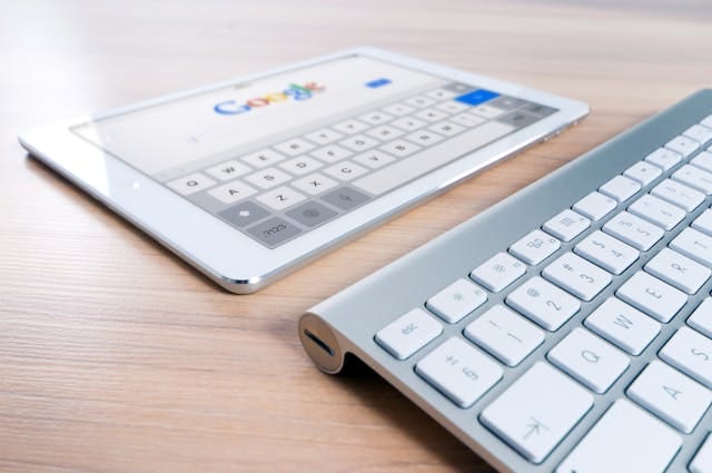 Na zdjęciu widoczne są tablet z otwartą wyszukiwarką Google i klawiatura, leżące na drewnianym biurku. Grafika odzwierciedla początkowy etap poszukiwań i researchu, który prowadzi do ustalenia cennika gotowych tekstów na strony internetowe