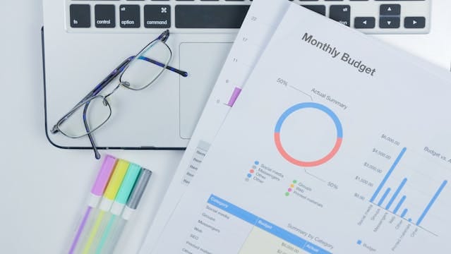 Zdjęcie z góry przedstawia arkusz kalkulacyjny z wykresem i napisem „Monthly Budget”. Obok leży laptop i okulary. Ilustracja pokazuje, jak ważne jest planowanie finansów, gdy rozważa ile kosztują teksty na stronę internetową czy jaki jest koszt prowadzenia bloga przez specjalistów z firmy Contentologia