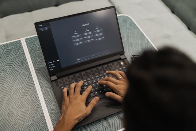 Ujęcie zza ramienia na osobę piszącą na klawiaturze laptopa, na którego ekranie włączona jest aplikacja ChatGPT z opcjami sugerowanych zapytań. Zdjęcie może sugerować pytanie, czy warto używać AI do treści marketingowych?