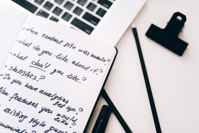 Zdjęcie przedstawia notatnik z ręcznie pisanymi pytaniami (m.in. "what do you like about it?", "Statistics?", "Do you have analytics set up?"). Notatnik leży na białym biurku obok laptopa, czarnego markera i spinacza do papieru. Wizualizuje to proces rozpoczynający współpracę, w którym agencje marketingowe definiują zakres usług i tworzą strategię opartą na dogłębnej analizie.