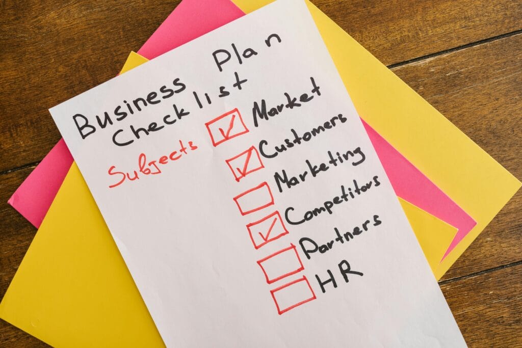 Ręcznie napisana Business Checklista (lista kontrolna biznesowa) na białej kartce, położonej na stosie kolorowych kartek. Lista zawiera pozycje do sprawdzenia, takie jak: Market, Customers, Marketing, Competitors, Partners, HR. Pozycje Market, Customers i Competitors są już zaznaczone czerwonymi ptaszkami. Reprezentuje to kluczowe obszary do weryfikacji doświadczenia i planu działania, który świadczą agencje marketingowe.