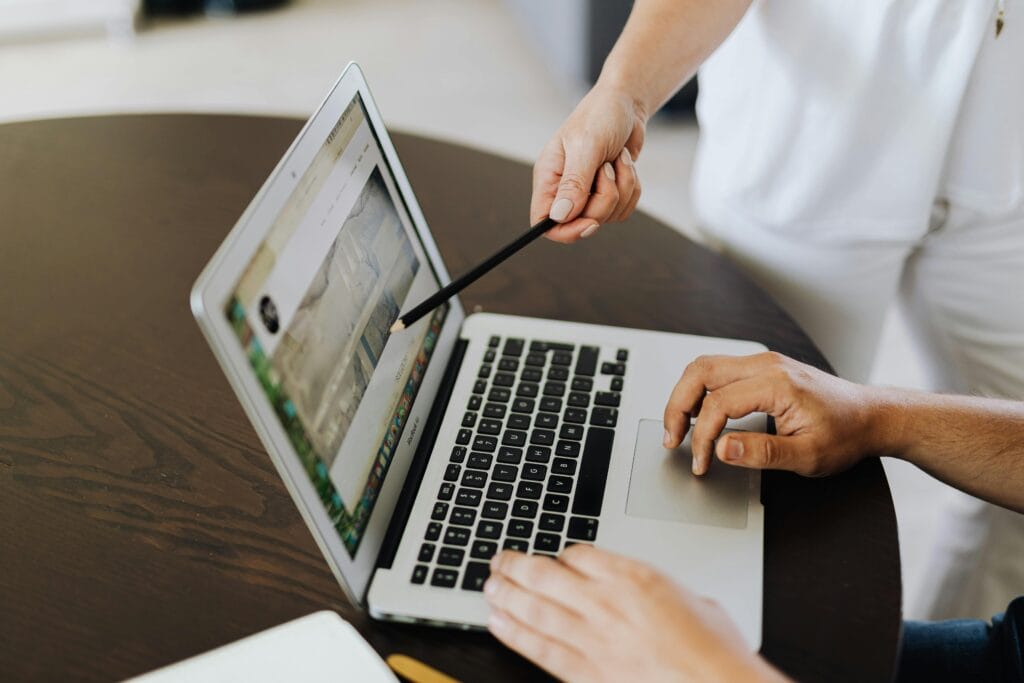 Dwie osoby pracujące przy laptopie na drewnianym stole: jedna wskazuje ołówkiem na ekran, na którym widoczna jest strona internetowa lub projekt graficzny. Obraz symbolizuje prezentację portfolio i omówienie case studies, co jest podstawą weryfikacji kompetencji, zanim wybierze się agencje marketingowe.