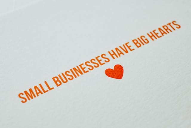 Zbliżenie na fragment białej kartki z ukośnym, pomarańczowym nadrukiem: "SMALL BUSINESSES HAVE BIG HEARTS" (Małe firmy mają duże serca), obok którego znajduje się czerwone, wypukłe serce. Podkreśla to ideę, że mniejsza, wyspecjalizowane agencja content marketingowa Contentologia oferuje bardziej spersonalizowane i zaangażowane podejście do klienta.