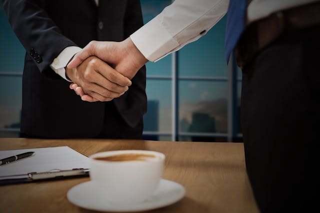 Bliskie ujęcie na uścisk dłoni dwóch biznesmenów w garniturach nad drewnianym stołem z kubkiem kawy i dokumentami, na tle dużego okna. Ilustruje to finalizację udanych negocjacji, do których prowadzi SEO marketing B2B w firmie Contentologia