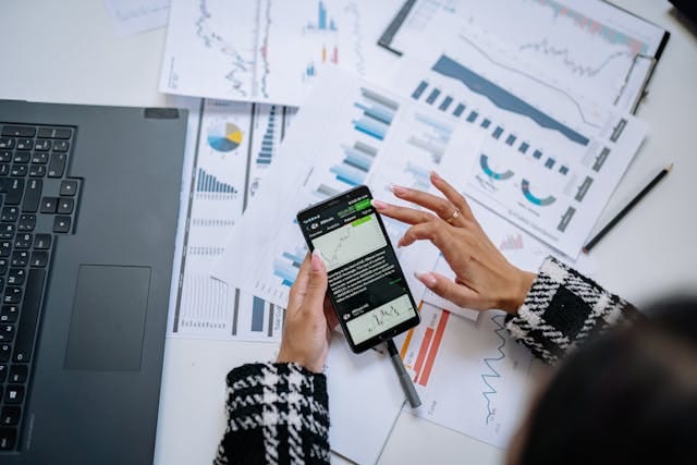 Ujęcie z góry na dłonie trzymające smartfona z aplikacją do analizy finansowej, otoczone wydrukami wykresów i laptopem. W dobie mobile-first, optymalizacja pod urządzenia mobilne jest kluczowa w Strategia SEO w B2B. Skorzystanie z usług profesjonalnej agencji marketingowej Contetnologia pozwala uniknąć kosztownych błędów samodzielnego tworzenia strategii seo dla B2B.