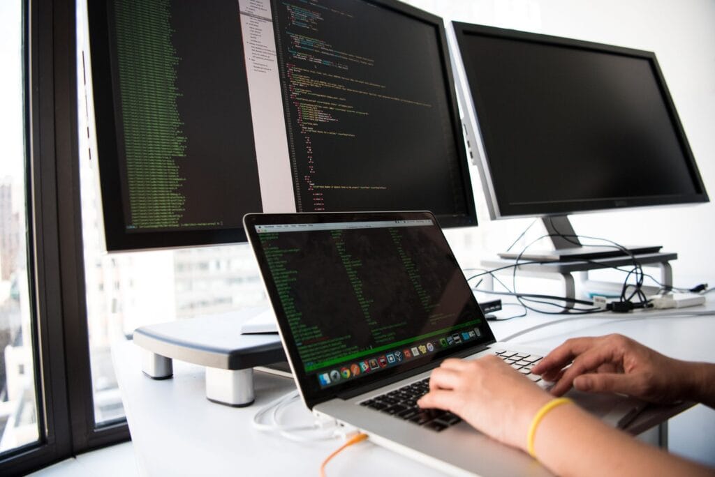 Zbliżenie na dłonie programisty intensywnie piszącego kod na laptopie, wspieranym przez dwa zewnętrzne monitory wyświetlające logi. Taka atmosfera pracy jest punktem wyjścia dla naszych analiz w Contentologii, gdzie przekładamy trudne wyzwania technologiczne na zrozumiałe case study, wspierając tym samym procesy sprzedażowe naszych klientów z branży technologicznej.