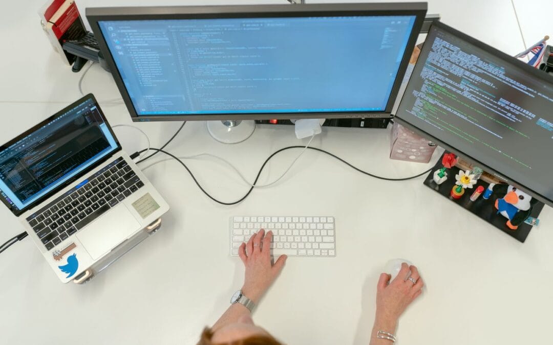 Widok z góry na stanowisko pracy programisty z trzema ekranami, na których wyświetlany jest złożony kod źródłowy. Ta profesjonalna przestrzeń odzwierciedla rygorystyczne podejście Contentologii do tworzenia treści, gdzie każdy tekst o architekturze technicznej jest precyzyjnie dopasowany do potrzeb Technical Validatora w ramach strategii content marketing IT.
