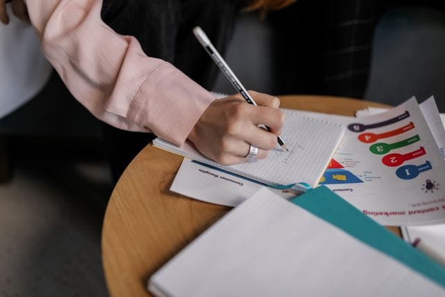 Zbliżenie na dłoń kobiety w jasnoróżowej koszuli, która pisze ołówkiem w notesie w kratkę, leżącym na okrągłym, drewnianym stoliku kawowym. Obok notesu ułożone są dokumenty i wydruki analityczne z kolorowymi wykresami przedstawiającymi etapy lejka sprzedażowego oraz widocznymi napisami, takimi jak „content marketing” oraz „consumer thoughts”. Cała kompozycja ukazuje proces planowania unikalnych działań promocyjnych, które na stronie Contentologia są fundamentem skutecznego docierania do klientów i budowania długofalowej widoczności organicznej w sieci.