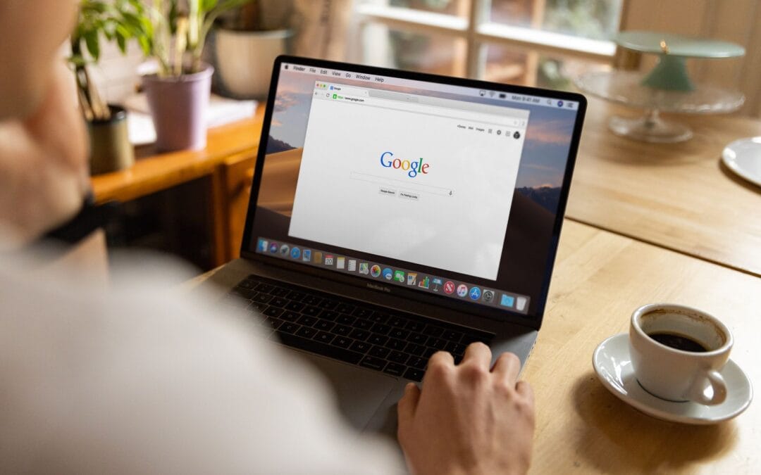 Zdjęcie przedstawiające perspektywę zza pleców osoby pracującej przy drewnianym biurku na laptopie MacBook Pro. Na jasnym ekranie komputera otwarta jest przeglądarka internetowa ze stroną główną wyszukiwarki Google. Prawa dłoń użytkownika spoczywa na obudowie laptopa w pobliżu klawiatury. Po prawej stronie, na jasnym blacie, stoi biała filiżanka ze spodkiem wypełniona czarną kawą, a w tle widać rozmyte wnętrze pomieszczenia z zielonymi roślinami doniczkowymi i oknem. Obraz ilustruje analizę patentów Google oraz wdrażanie strategii SEO przez agencję Contentologia.